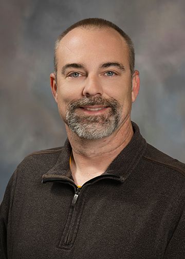 A man with a beard is wearing a brown sweater and smiling for the camera.