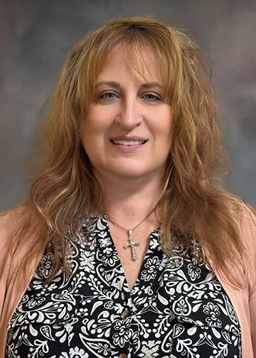 A woman wearing a floral shirt and a cross necklace is smiling for the camera.
