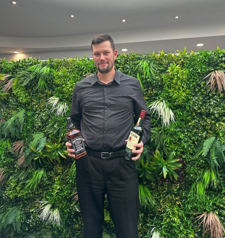 A man is holding two bottles of alcohol in front of a green wall.