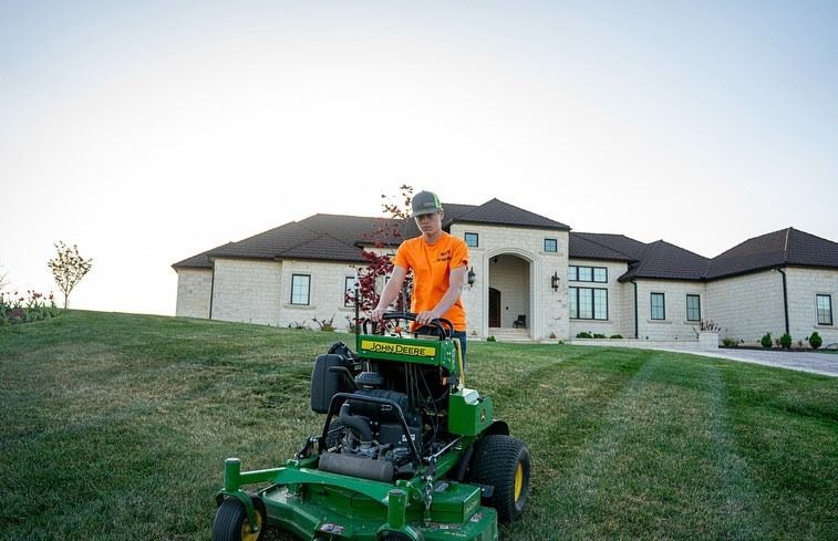 A person mowing a lawn with a green and yellow riding mower in front of a large house.