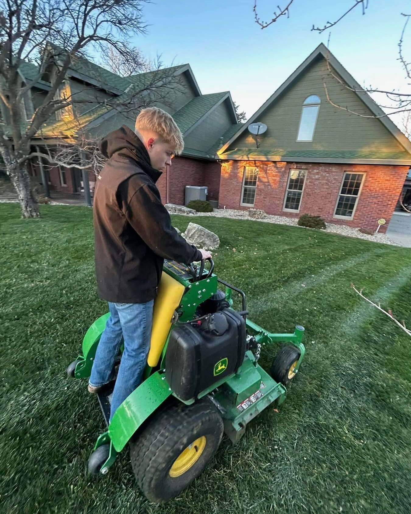 Young man mowing a green lawn on a John Deere riding mower in front of a brick and green-roofed house.