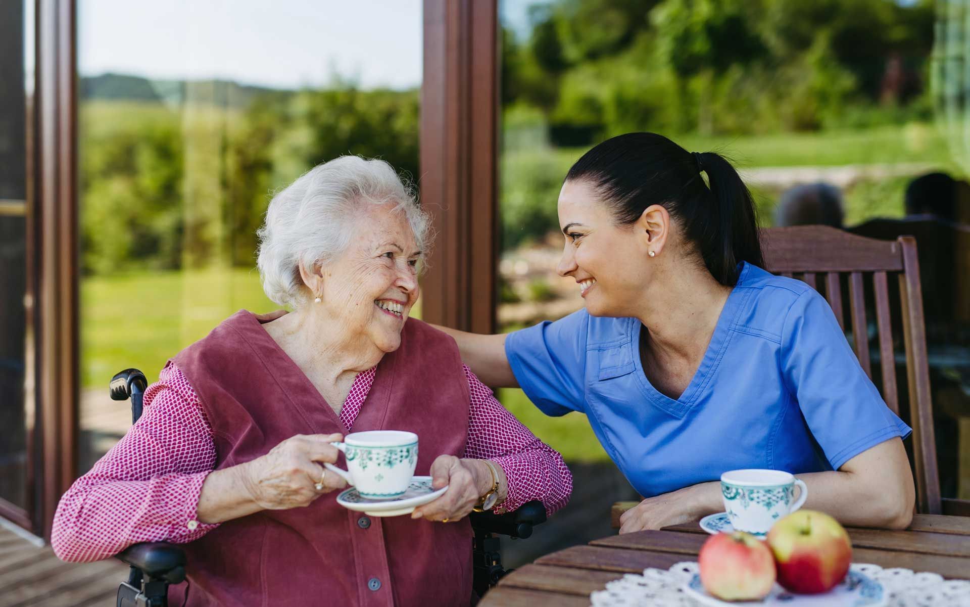 A senior caregiver is spending time with an elderly woman, drinking tea, and talking.