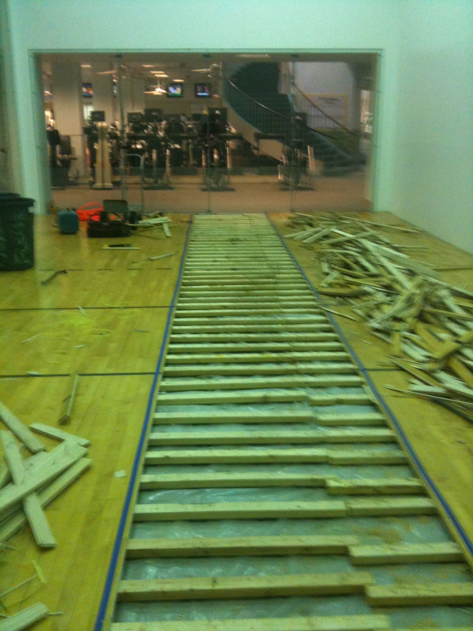 Long metal ladder laid on a gym floor, leading toward a glass-walled room.