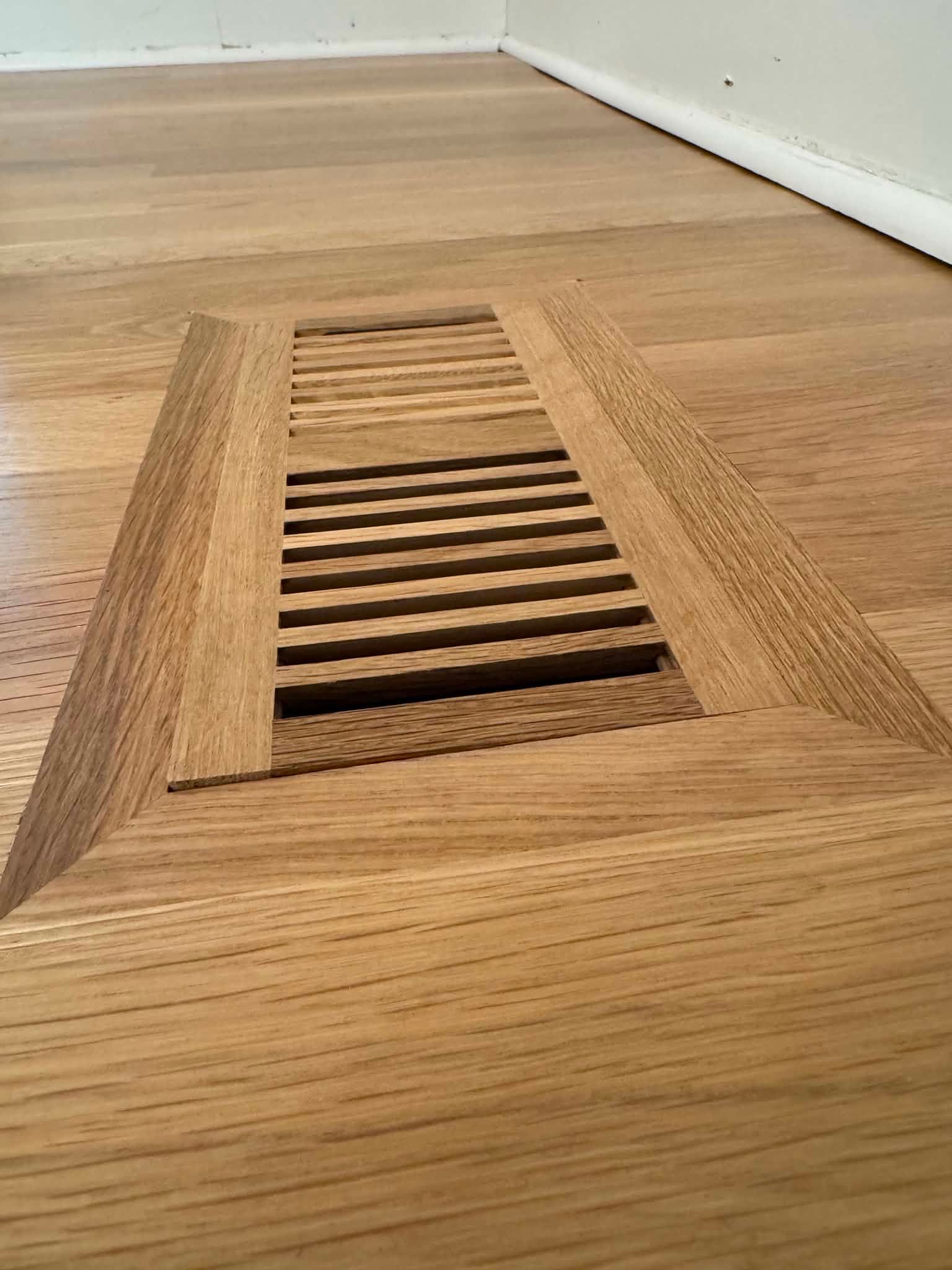 Wood floor vent grille inset in a light hardwood floor near a white baseboard.