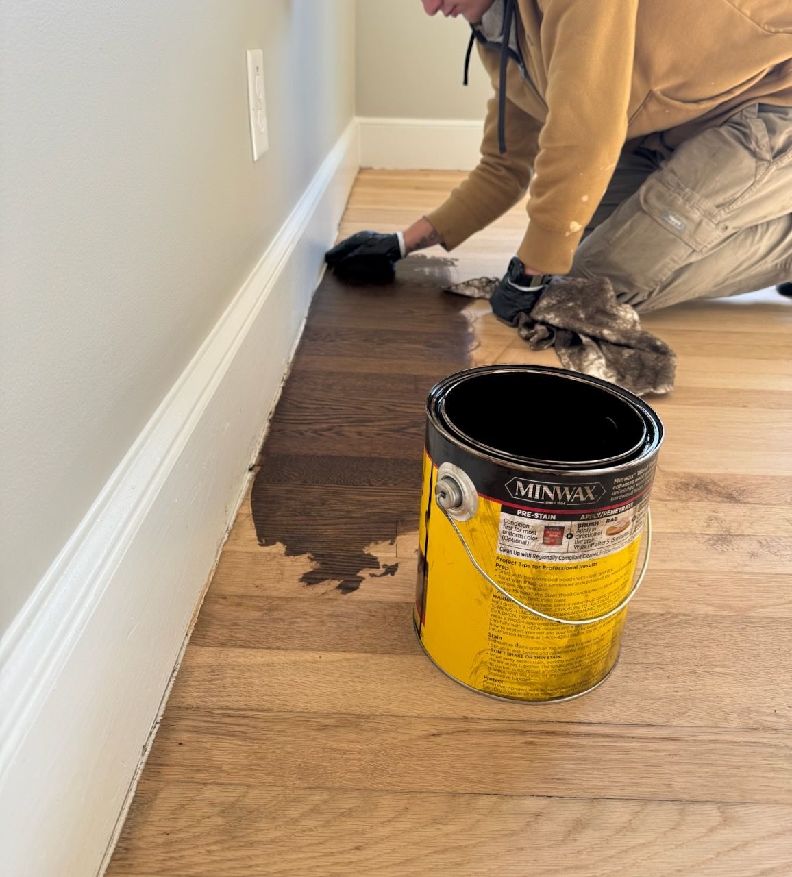 Person kneeling and painting a floor edge with a black tool beside a yellow paint can