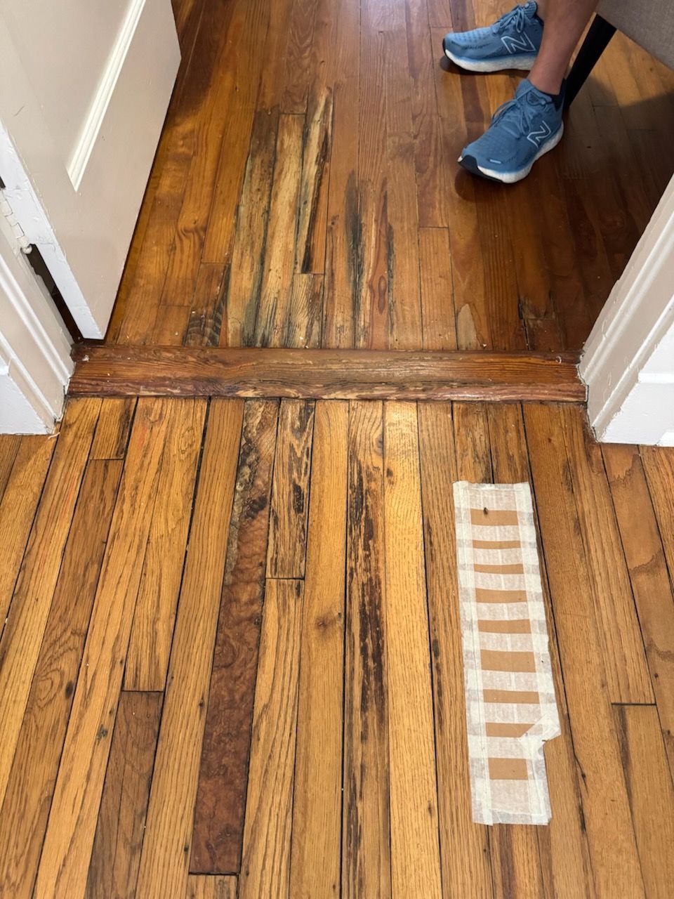 Worn wooden floor with a damaged threshold and a person's blue shoes at the top of the doorway