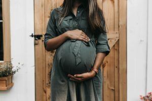 A pregnant woman is standing in front of a wooden door holding her belly.