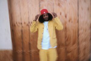 A man wearing a yellow suit and a red hat is standing in front of a wooden wall.