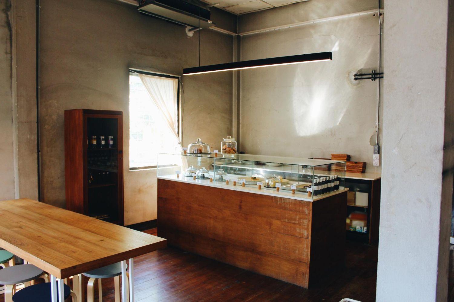 Cafe interior: wooden counter with pastries, wooden table, tall wooden cabinet, window, concrete walls.