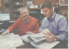 Jack and Jackson Bogan review documents at a desk in an office. One in red sweater smiles, other in blue shirt focuses.