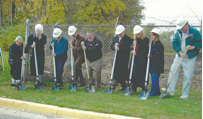Group of people in hard hats and jackets using shovels for a ground-breaking ceremony next to a chain-link fence.