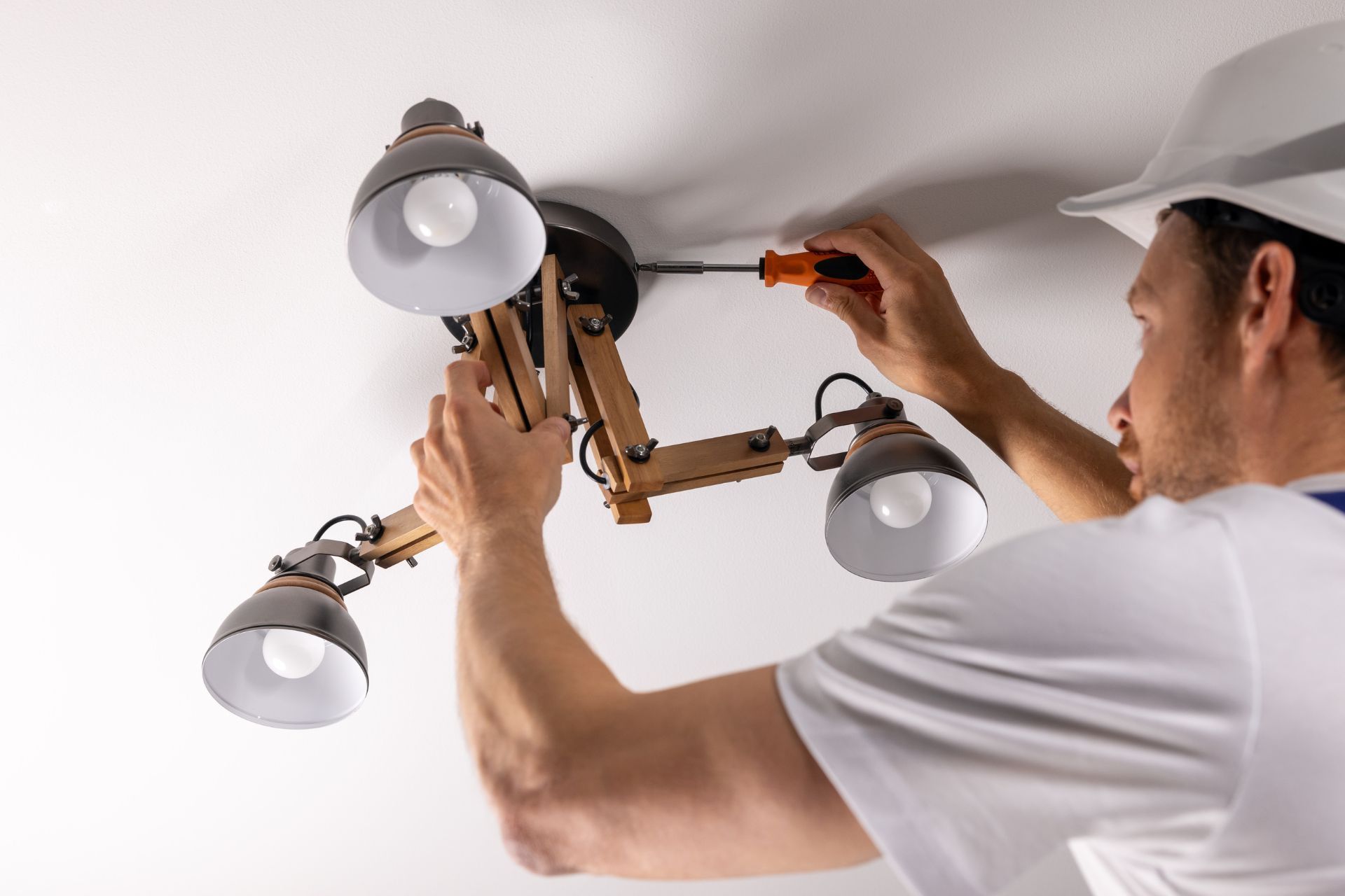 Person in a white shirt and hard hat installing a ceiling light fixture with a screwdriver.