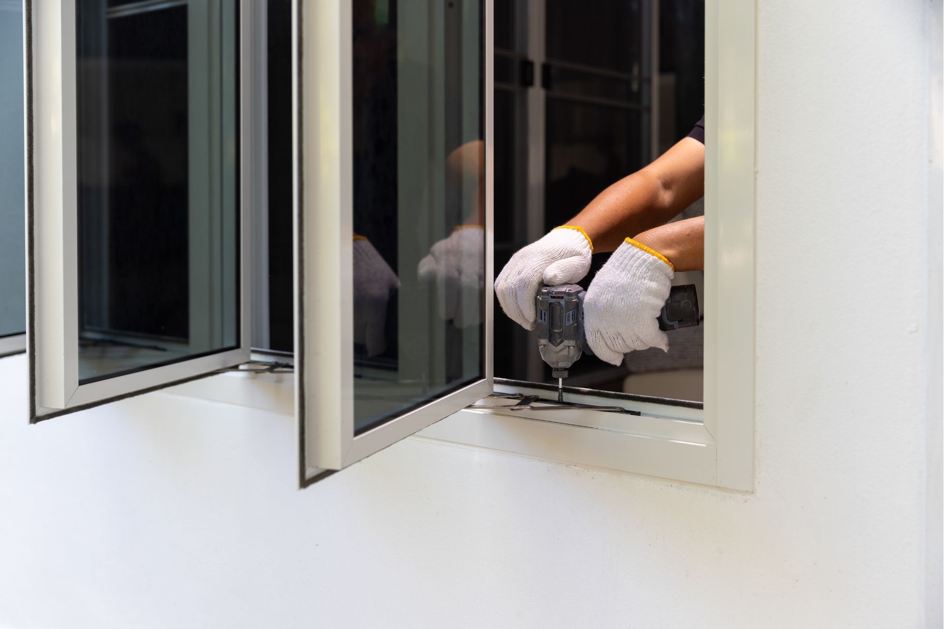 Person wearing gloves using a drill to install a window. White wall and open windows visible.