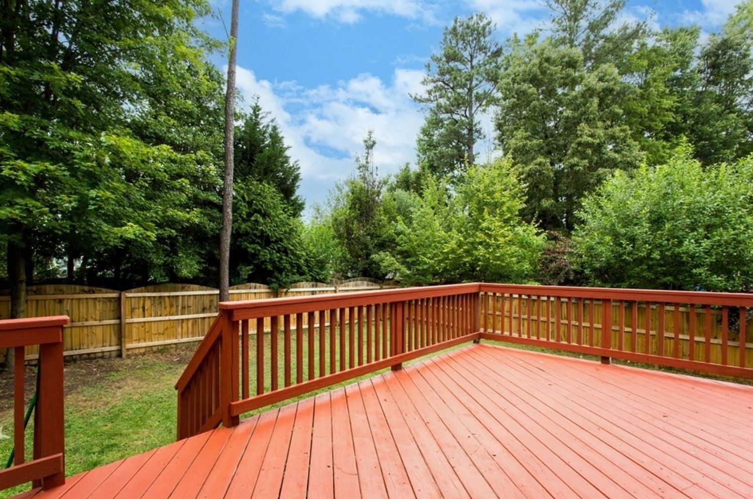 Red wooden deck overlooking a backyard with trees and a wooden fence under a blue sky.
