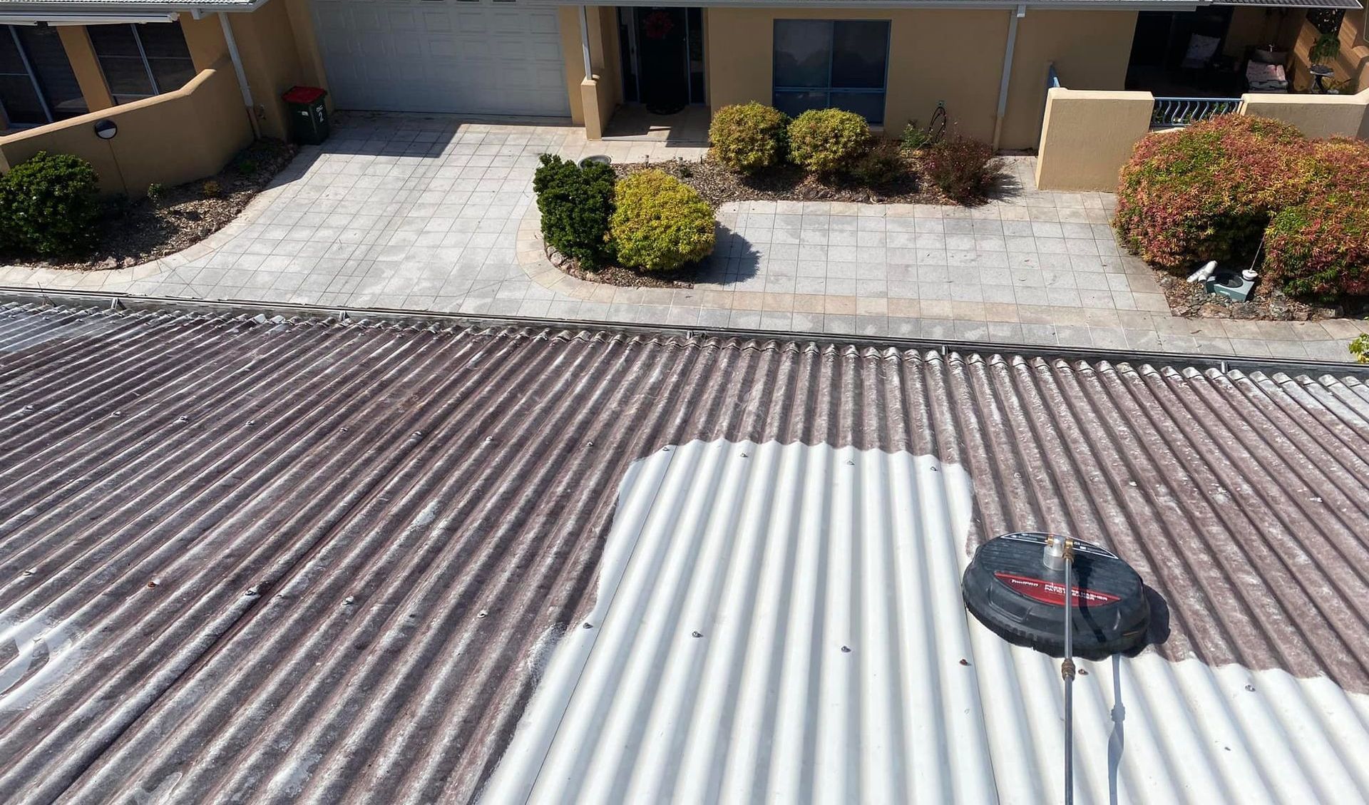 A Man Is Using A High Pressure Washer To Clean A Driveway In Front Of A House — The Guttering Cleaning Man in Forster, NSW
