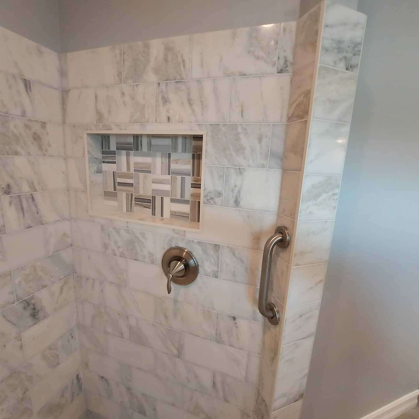 A marble-tiled shower wall features a mosaic-tiled recessed shelf, a metal shower handle, and a vertical grab bar.