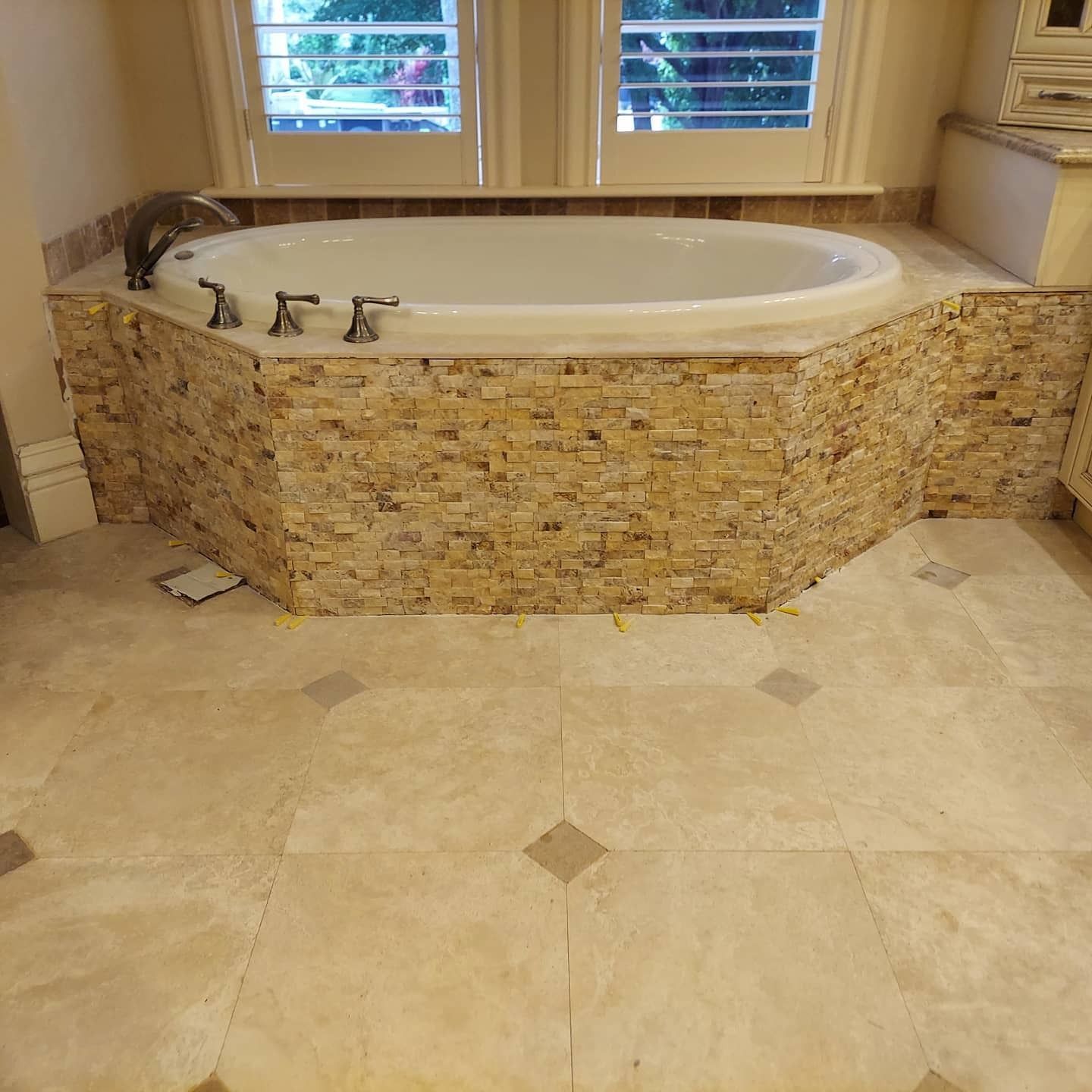 An oval soaking tub set in a deck finished with stacked stone, tiled with large light-colored floor tiles.