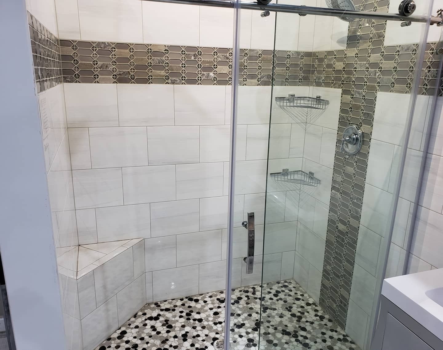 A modern shower enclosure with white tiled walls, a decorative stone border, a built-in seat, and a pebble-stone floor.