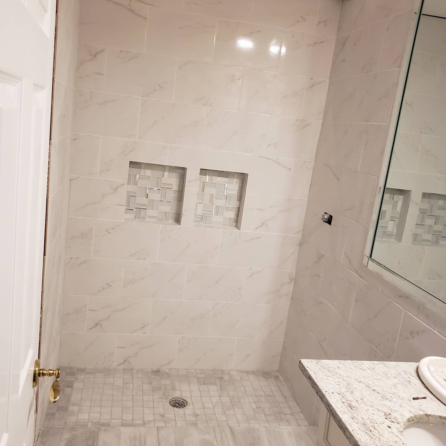 A walk-in shower with white marbled walls, mosaic tile niches, and a grey tiled floor, next to a marble bathroom vanity.