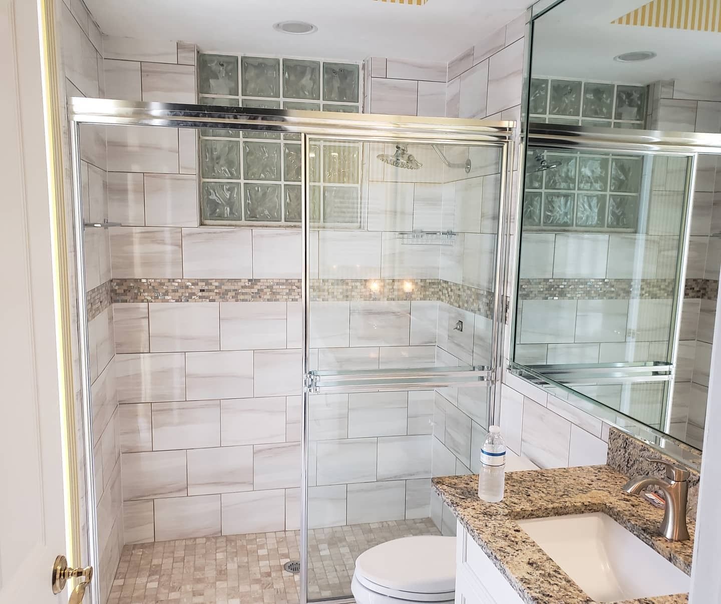 A bathroom featuring a glass shower with light-colored, textured stone-look tiles, a small window, and a granite vanity.