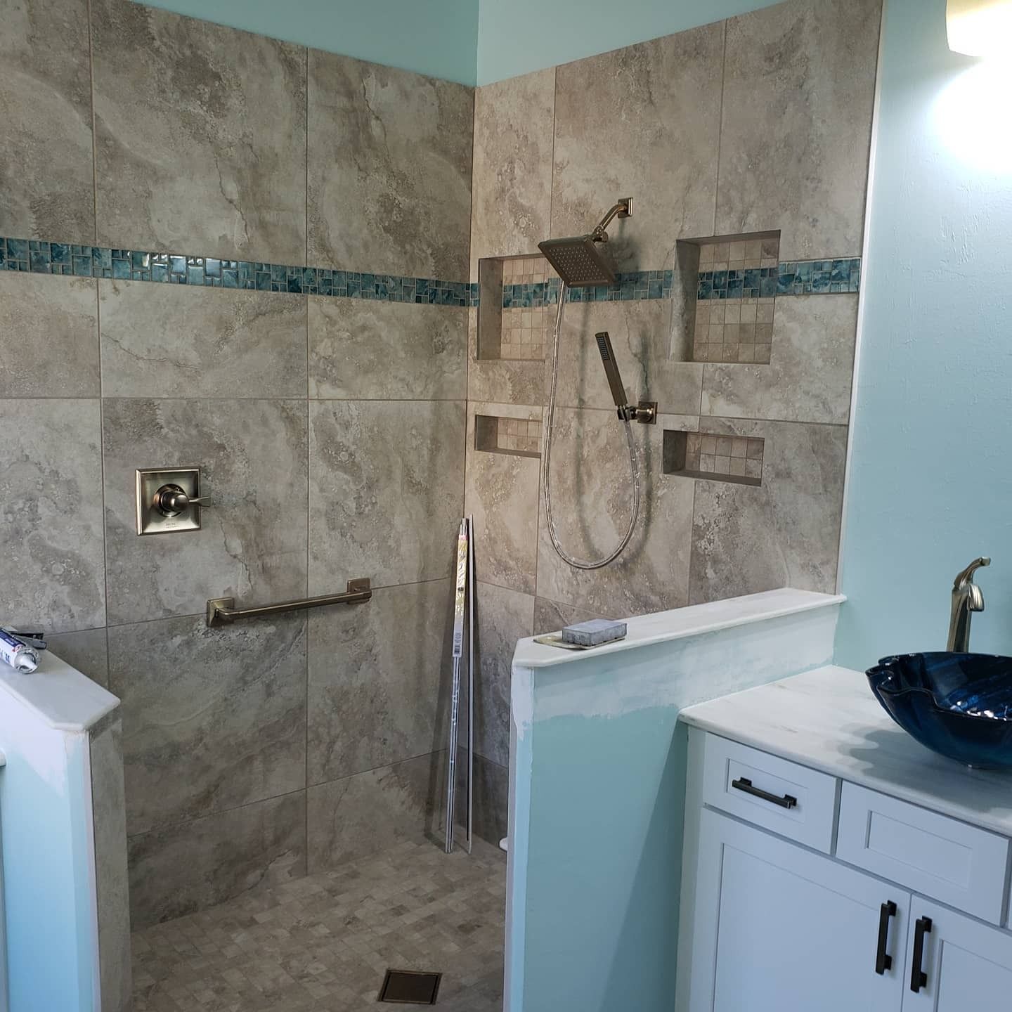A walk-in shower with gray stone tiles, a teal accent stripe, built-in niches, and a silver shower head next to a vanity.