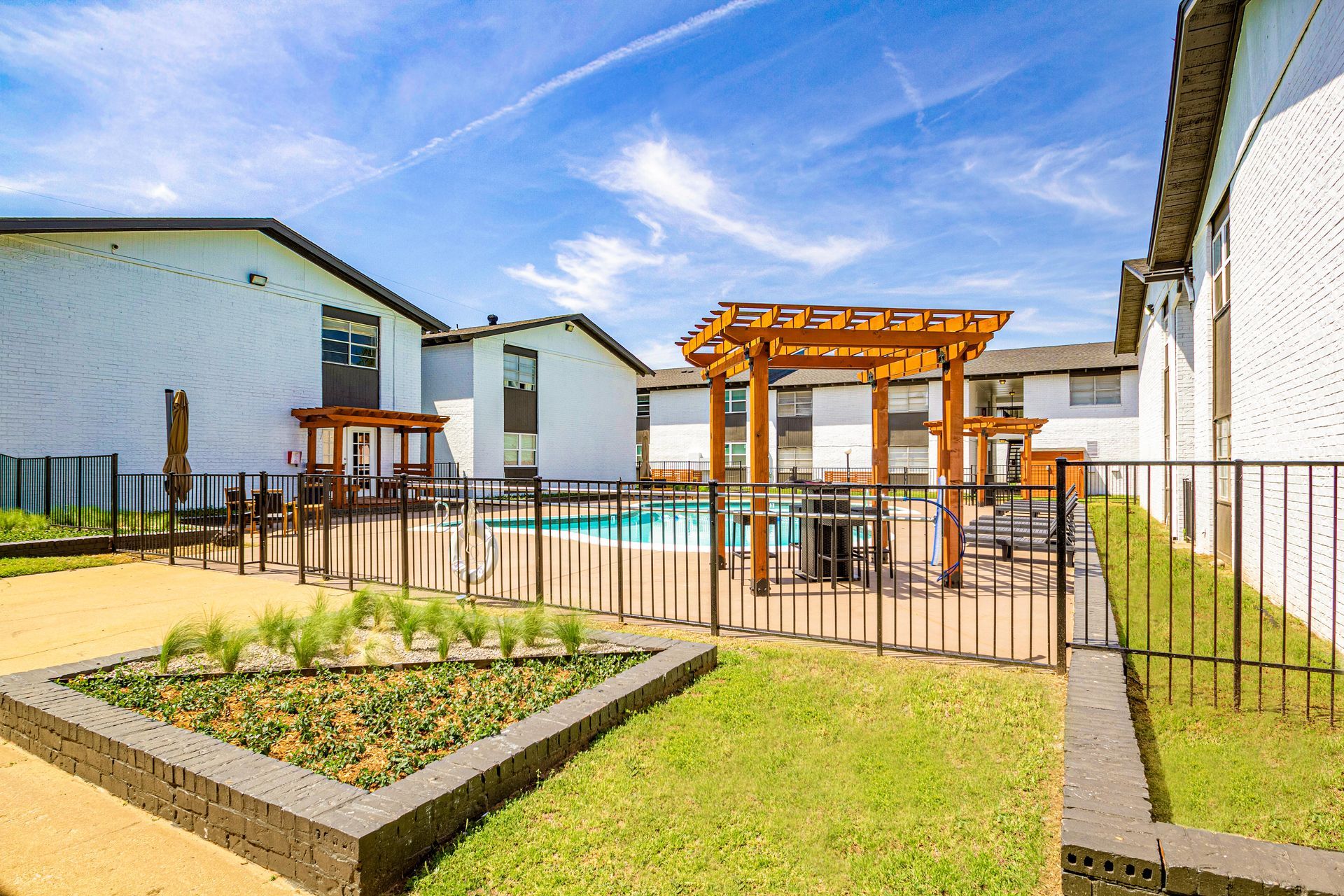 Pool area with pergola, fenced, between white buildings, against blue sky.