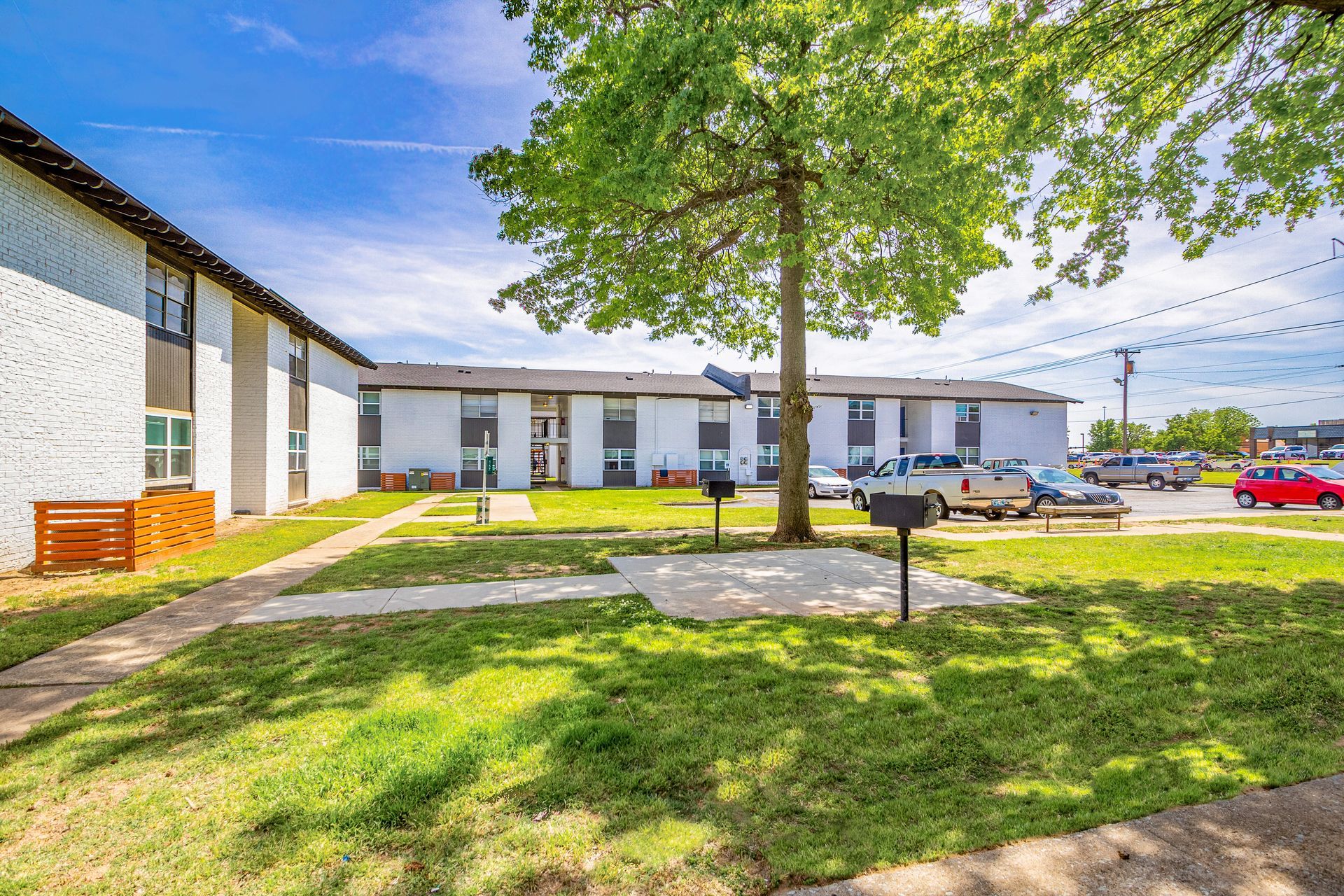 Exterior of a two-story apartment building with a grassy yard, trees, and parked cars under a blue sky.