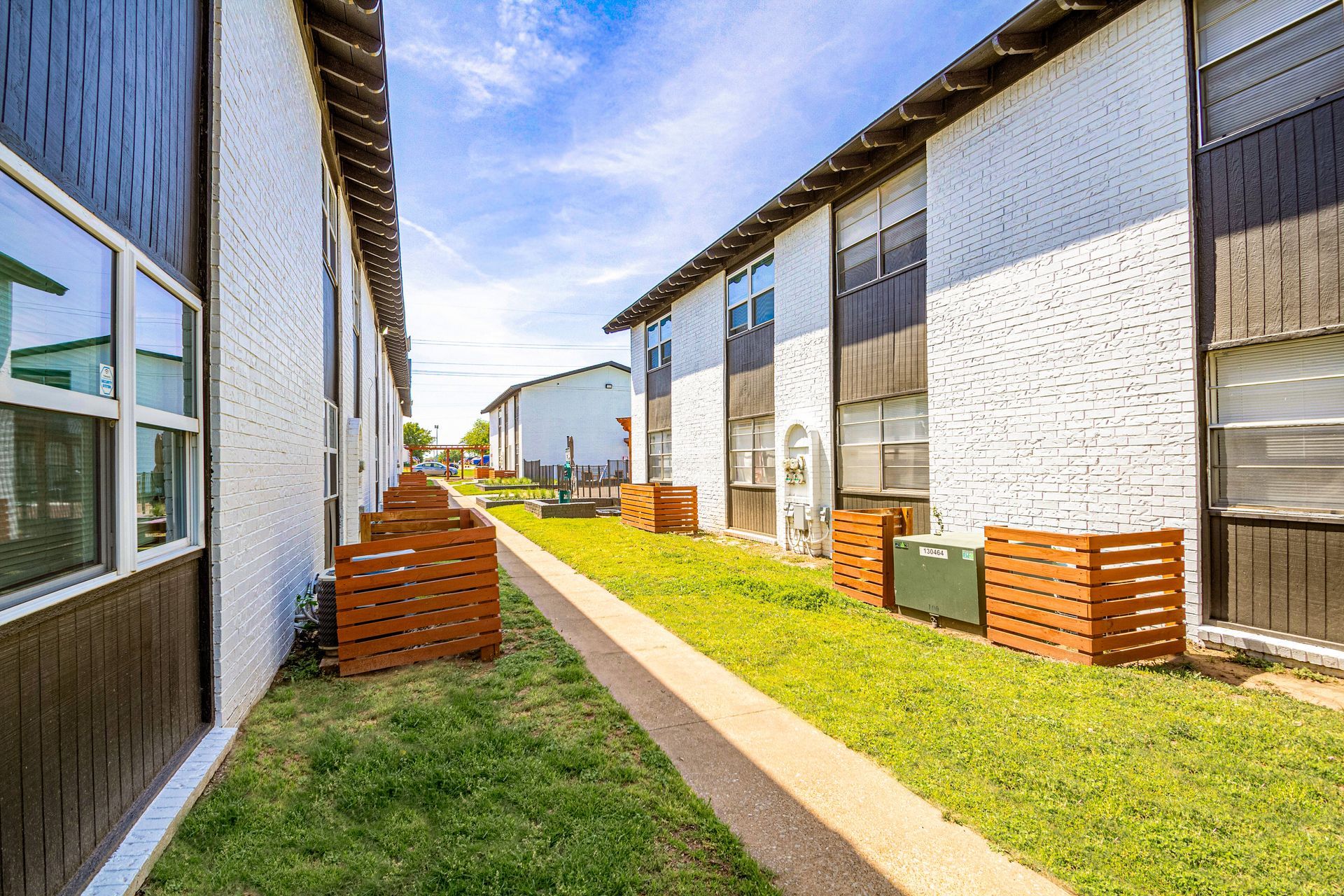 Apartment buildings with white brick walls, brown trim, and a grassy walkway. Sunny day.