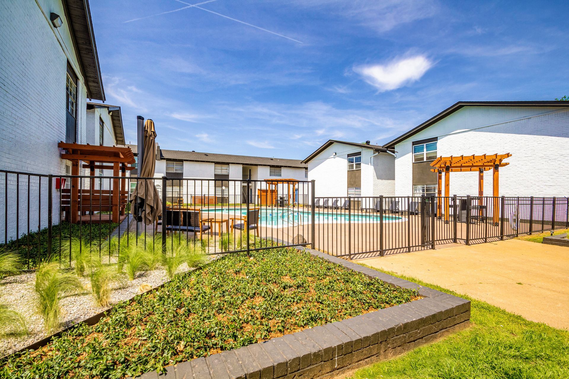 Courtyard with a pool, white buildings, wooden structures, and black fencing. Blue sky with clouds.