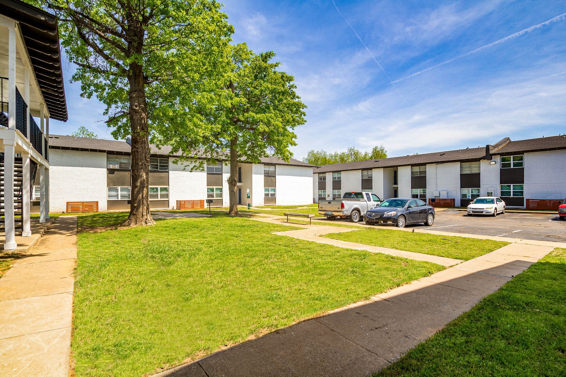 Apartment complex courtyard with cars parked, green lawn, trees, and clear blue sky.