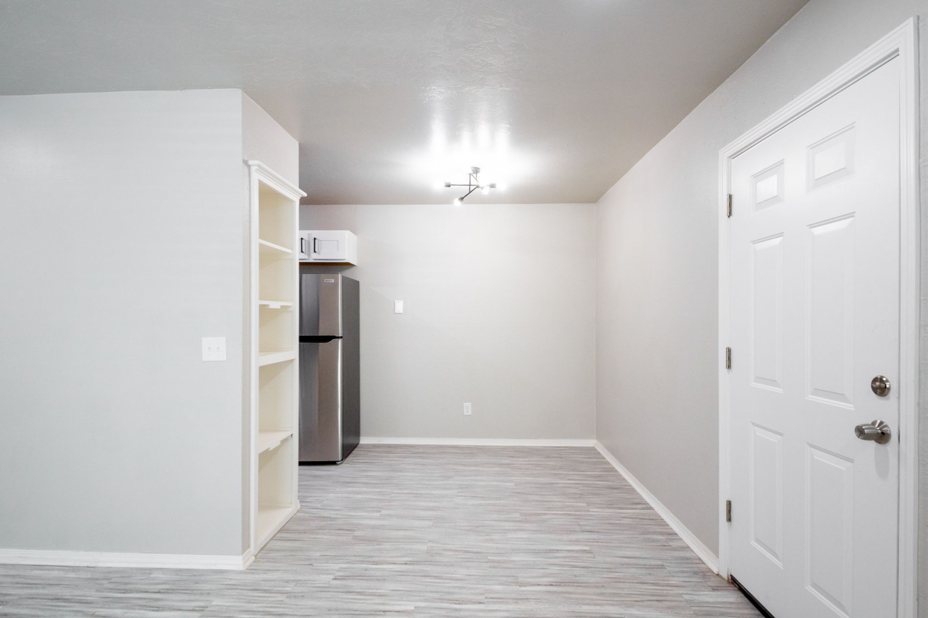 Interior of an apartment with gray walls, white door, light wood flooring, and a stainless steel refrigerator.