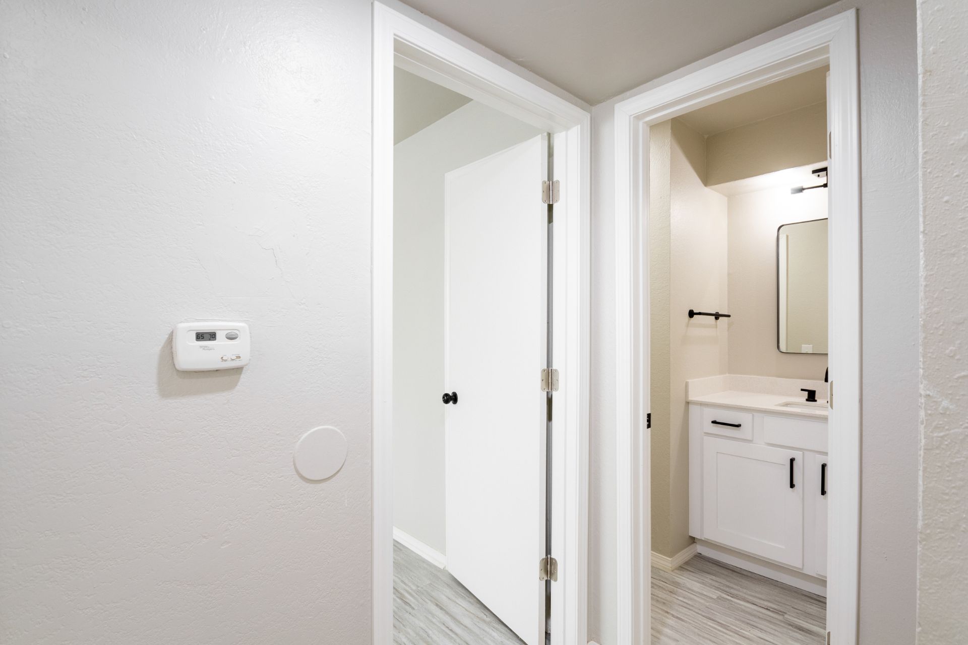Hallway with white walls, open doors to a bathroom and a room; thermostat on the left wall.