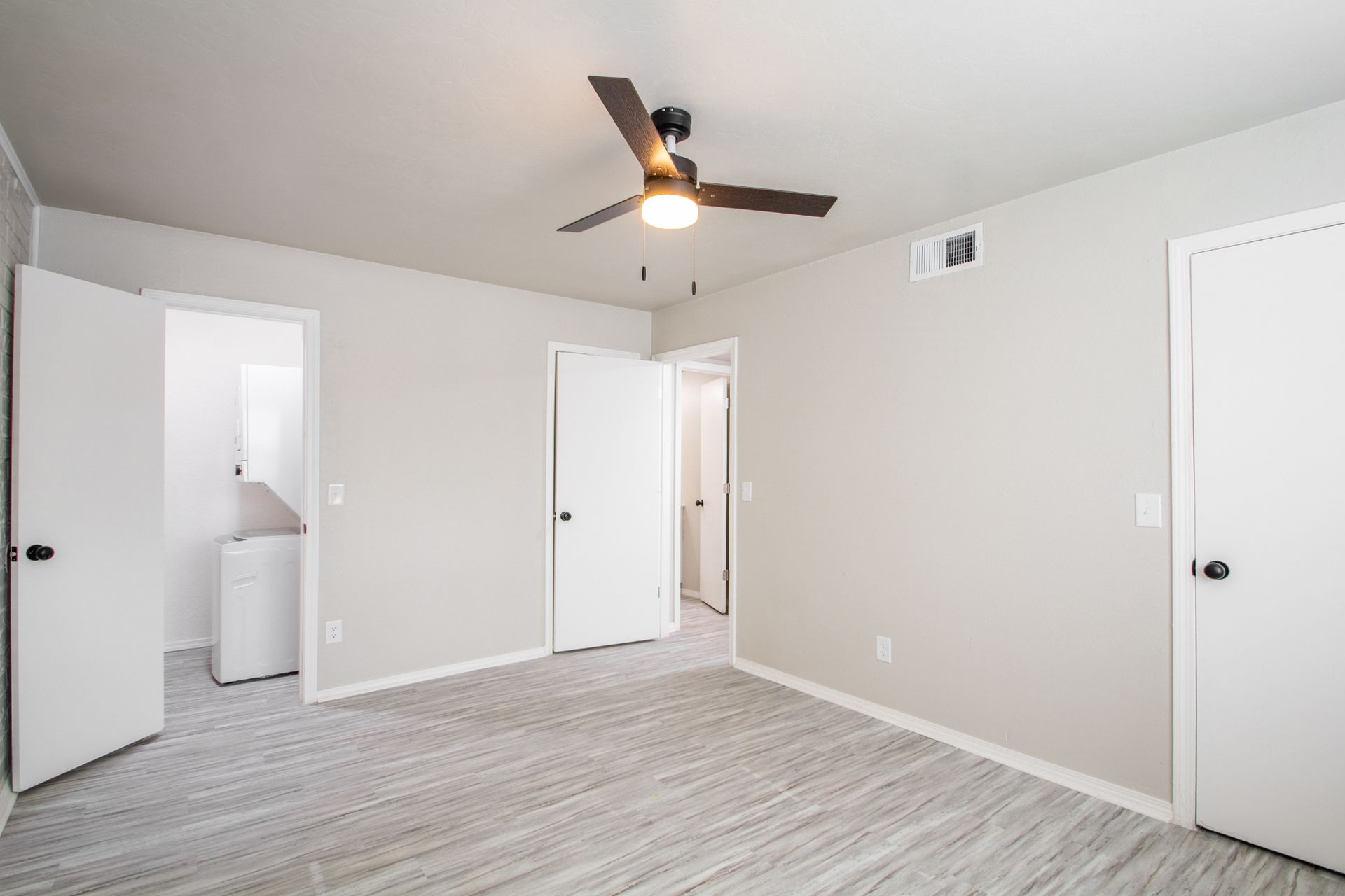 Empty bedroom with light gray walls and carpet, open doors to other rooms, ceiling fan.