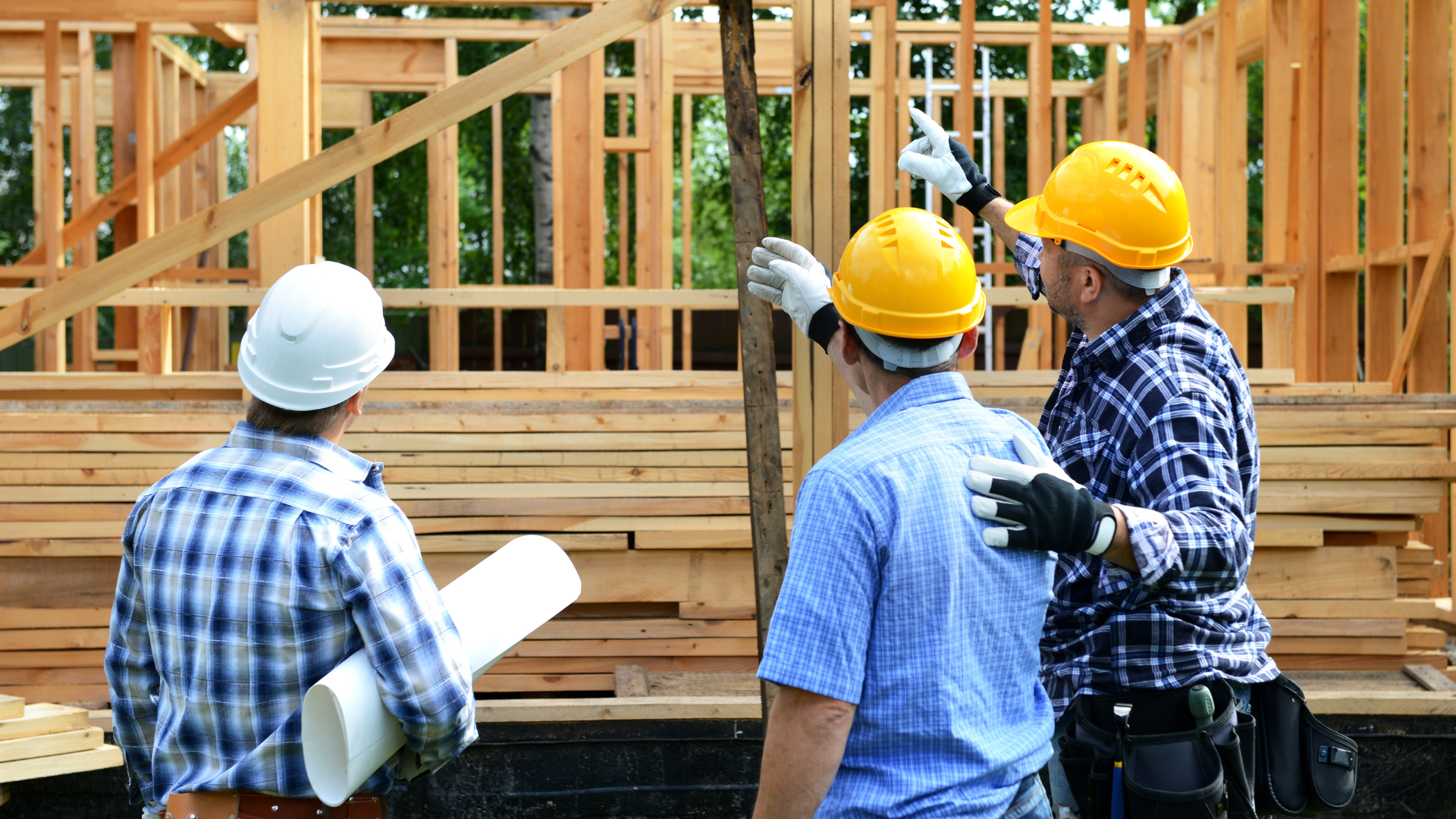 Three construction workers in hard hats examine a wooden house frame, one points upwards.