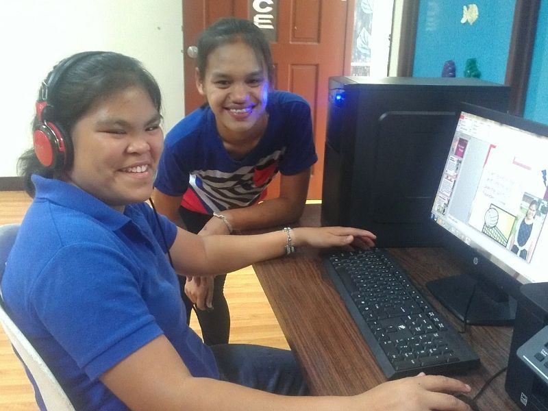 A girl wearing headphones sits next to another girl using a computer