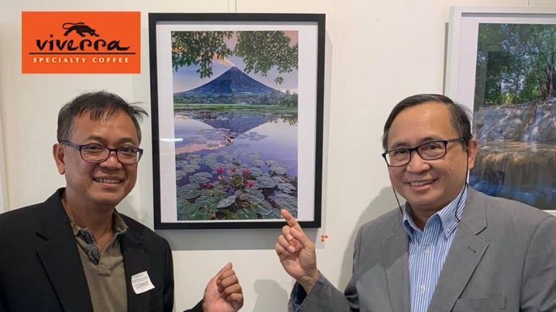 Two men are standing next to each other in front of a framed picture of a mountain.