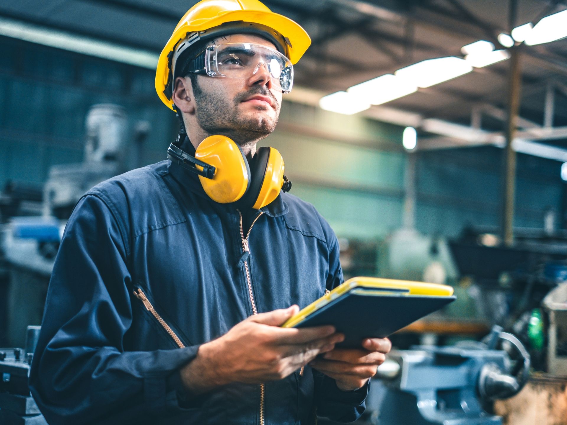 A man wearing a hard hat and safety glasses is holding a tablet in a factory.