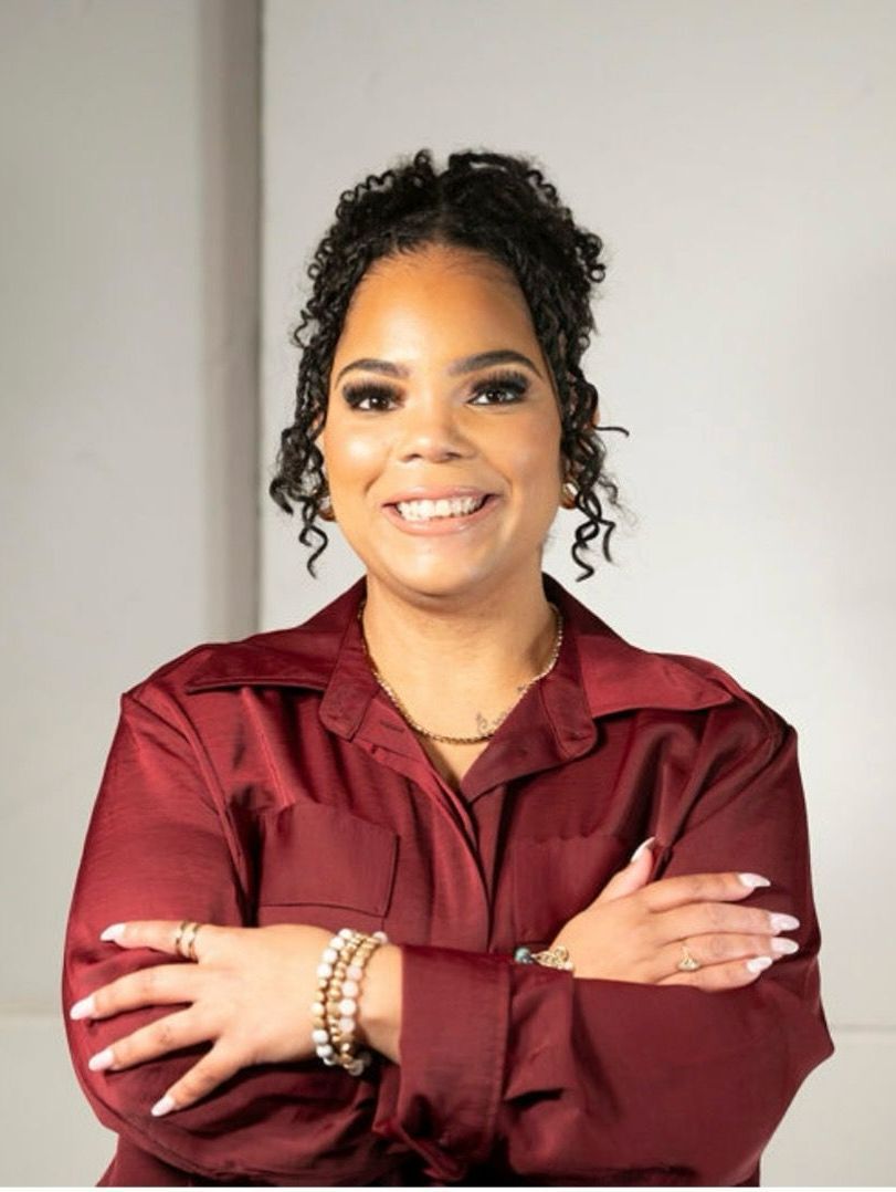 Woman with curly hair smiles, arms crossed, wearing a maroon shirt.