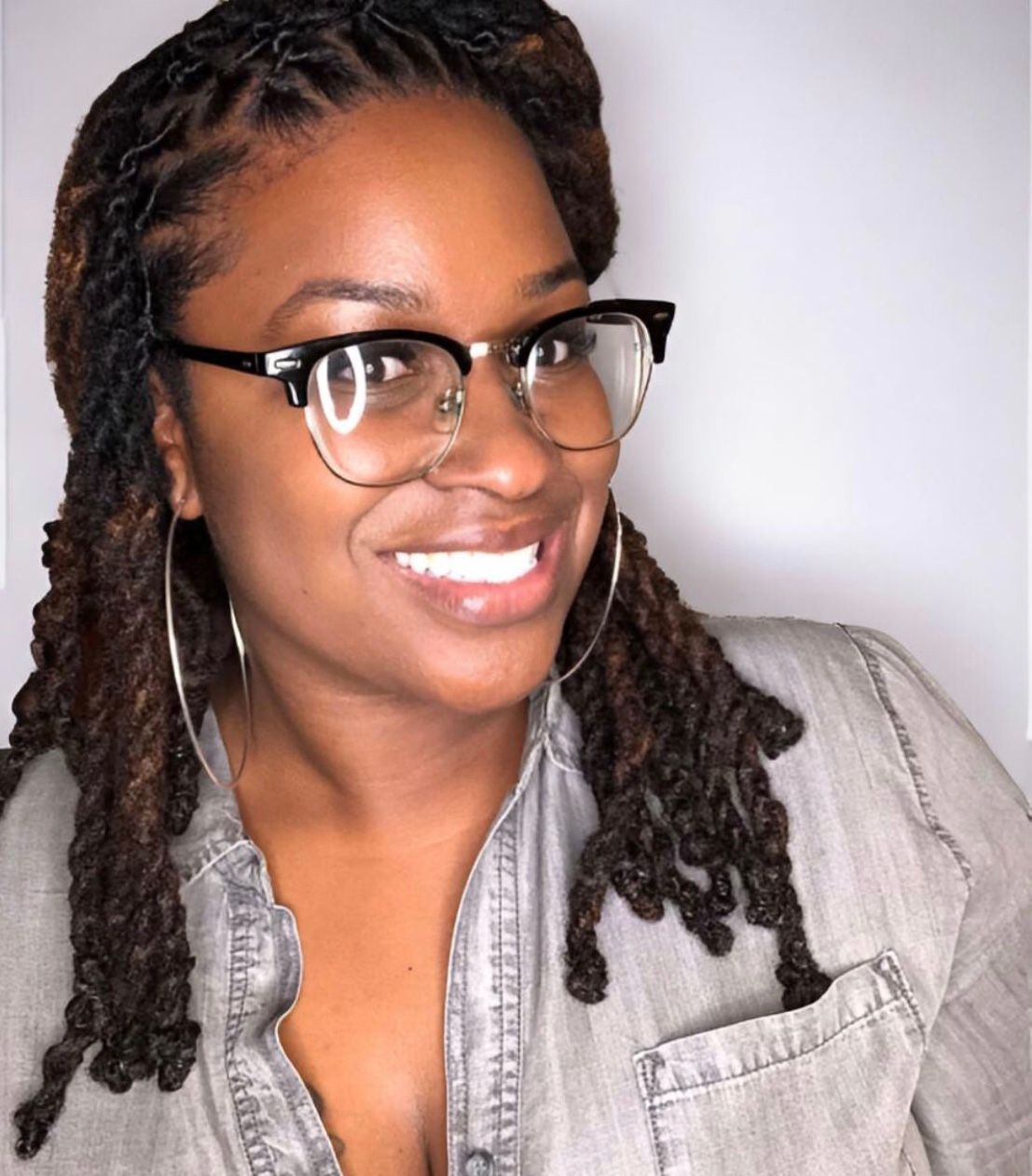 Woman with locs, glasses, and hoop earrings smiles; wearing a grey shirt.