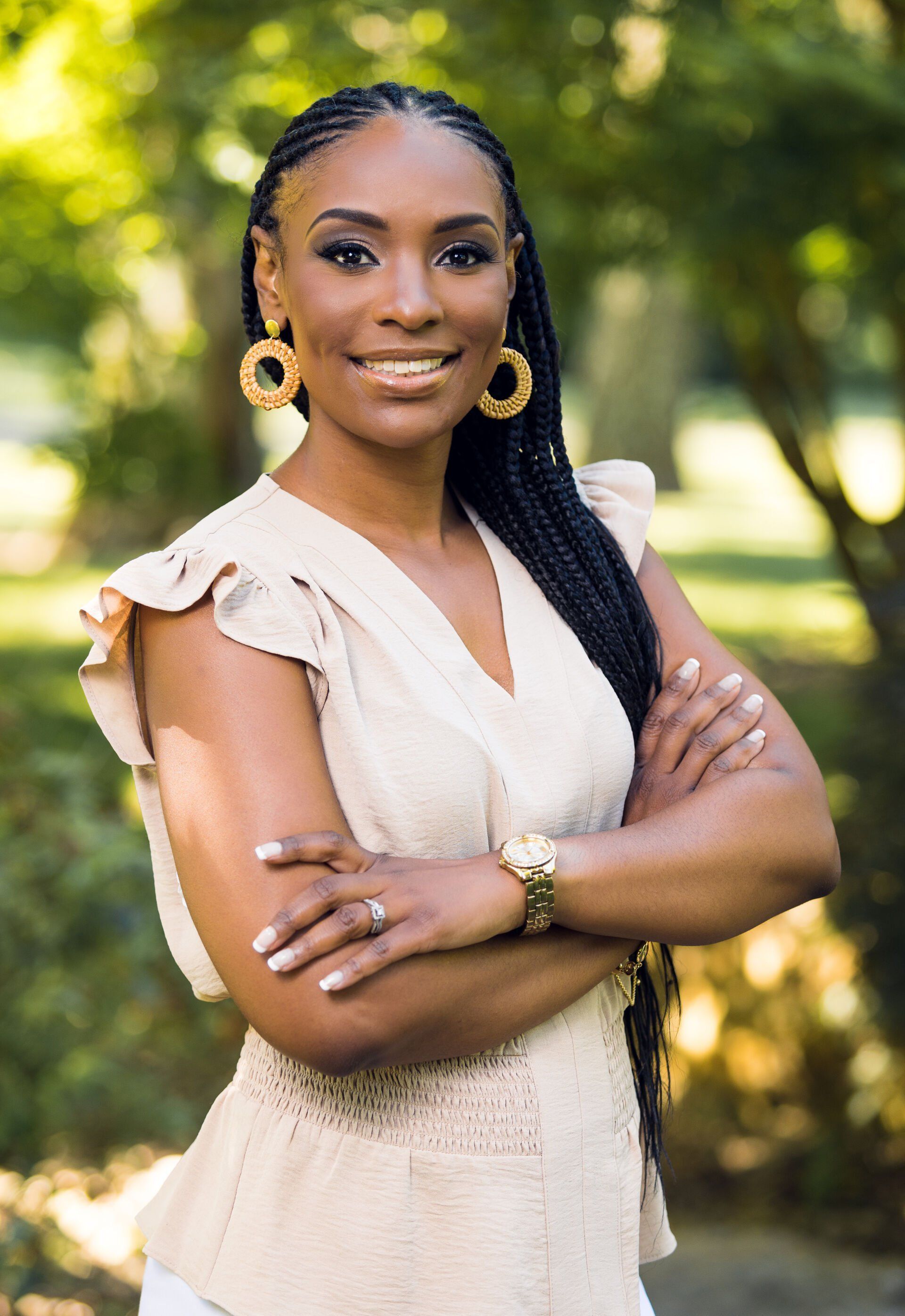 Woman with arms crossed, smiling, wearing tan top and gold earrings, outdoors in a park.