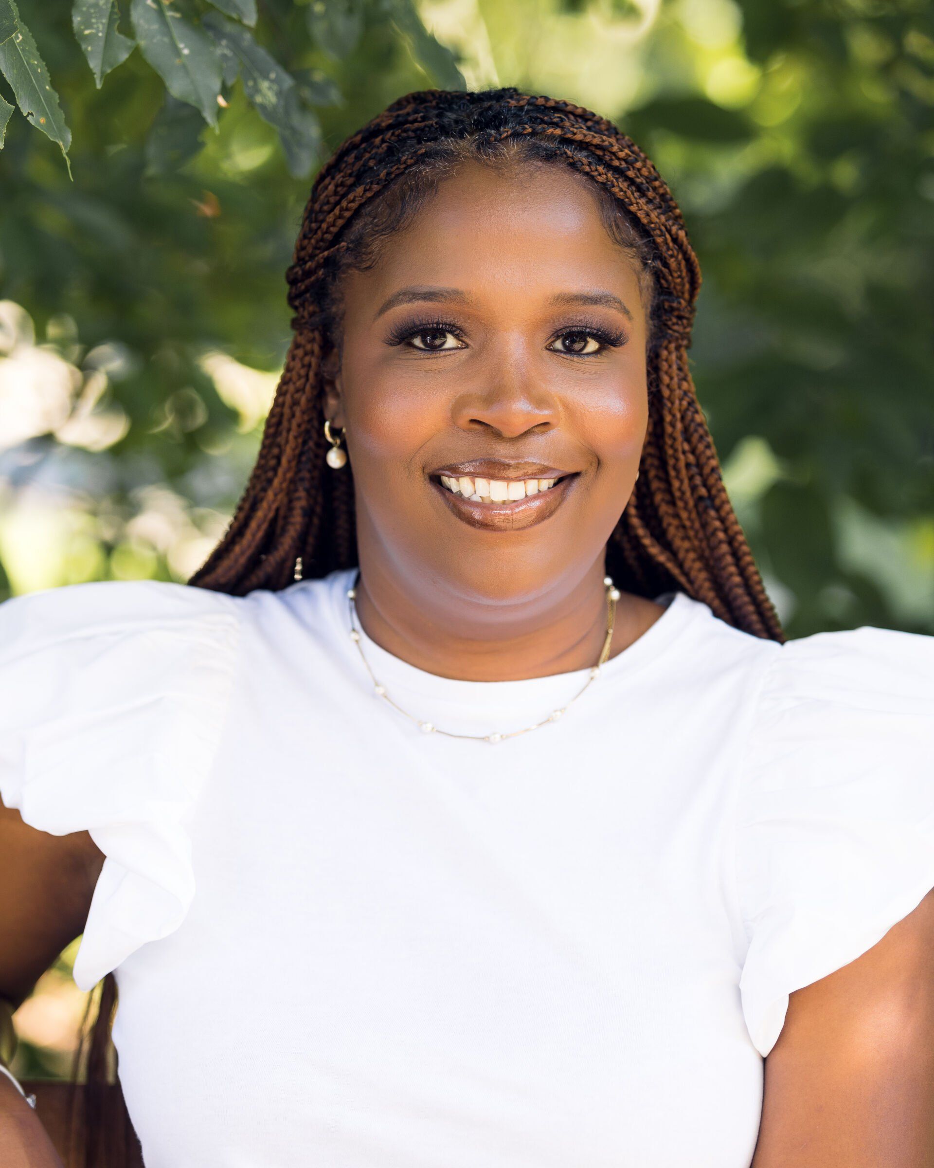 Woman with arms crossed, smiling, wearing tan top and gold earrings, outdoors in a park.