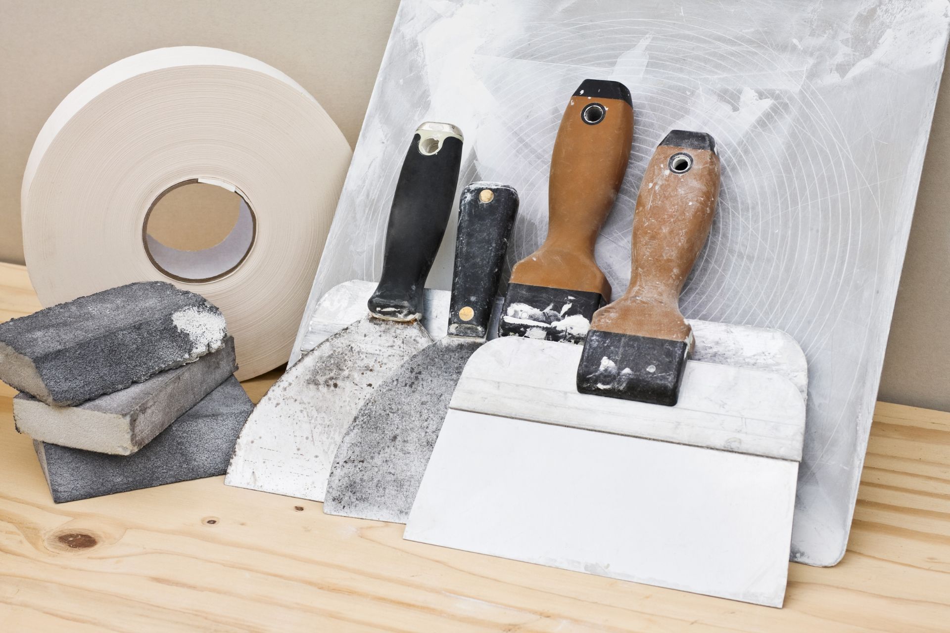 Drywall tools on a wooden surface: tape roll, sanding blocks, putty knives, and a taping knife.