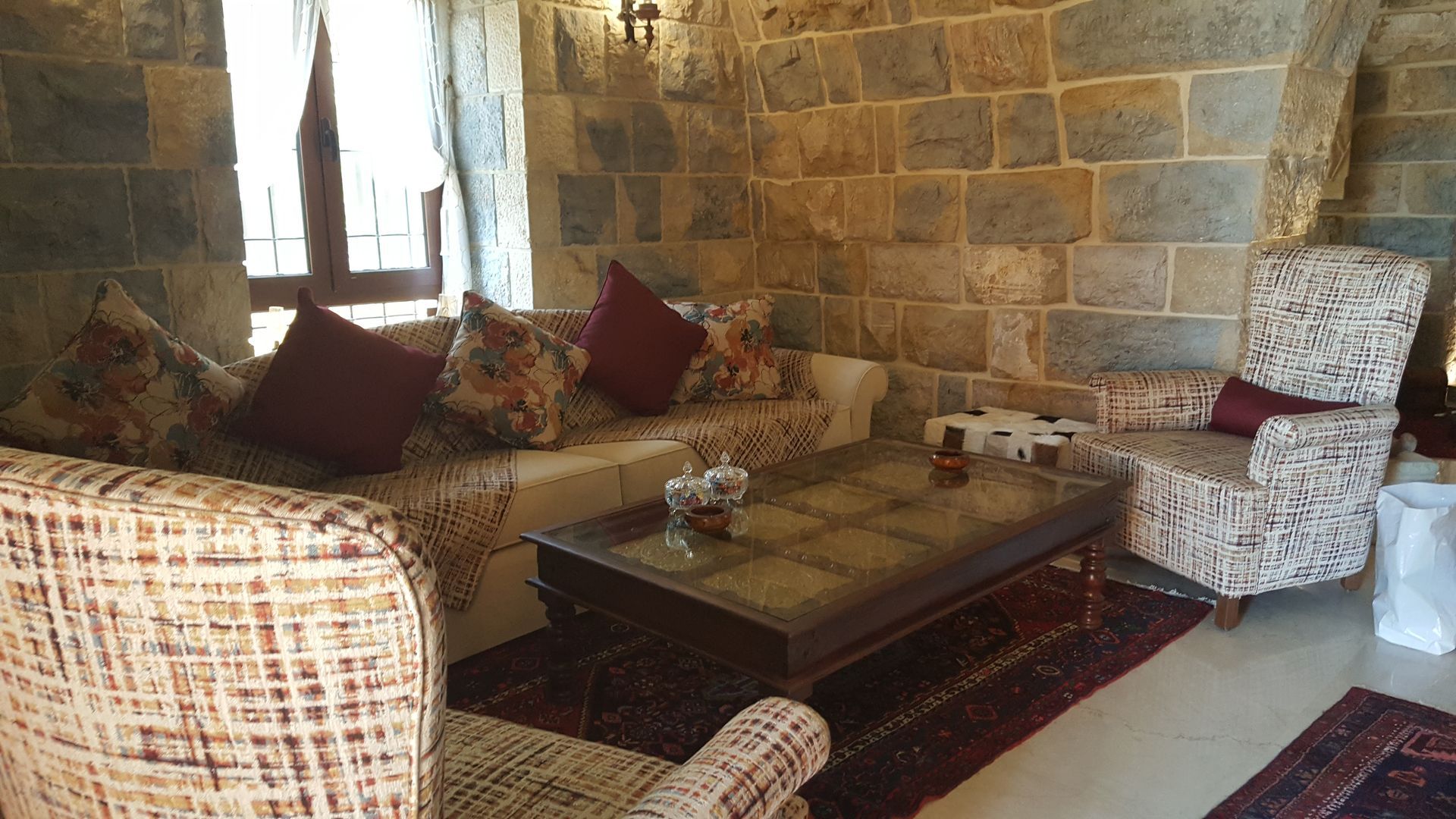 Cozy stone-walled living room with patterned furniture, a glass-topped coffee table, and small red pillows.