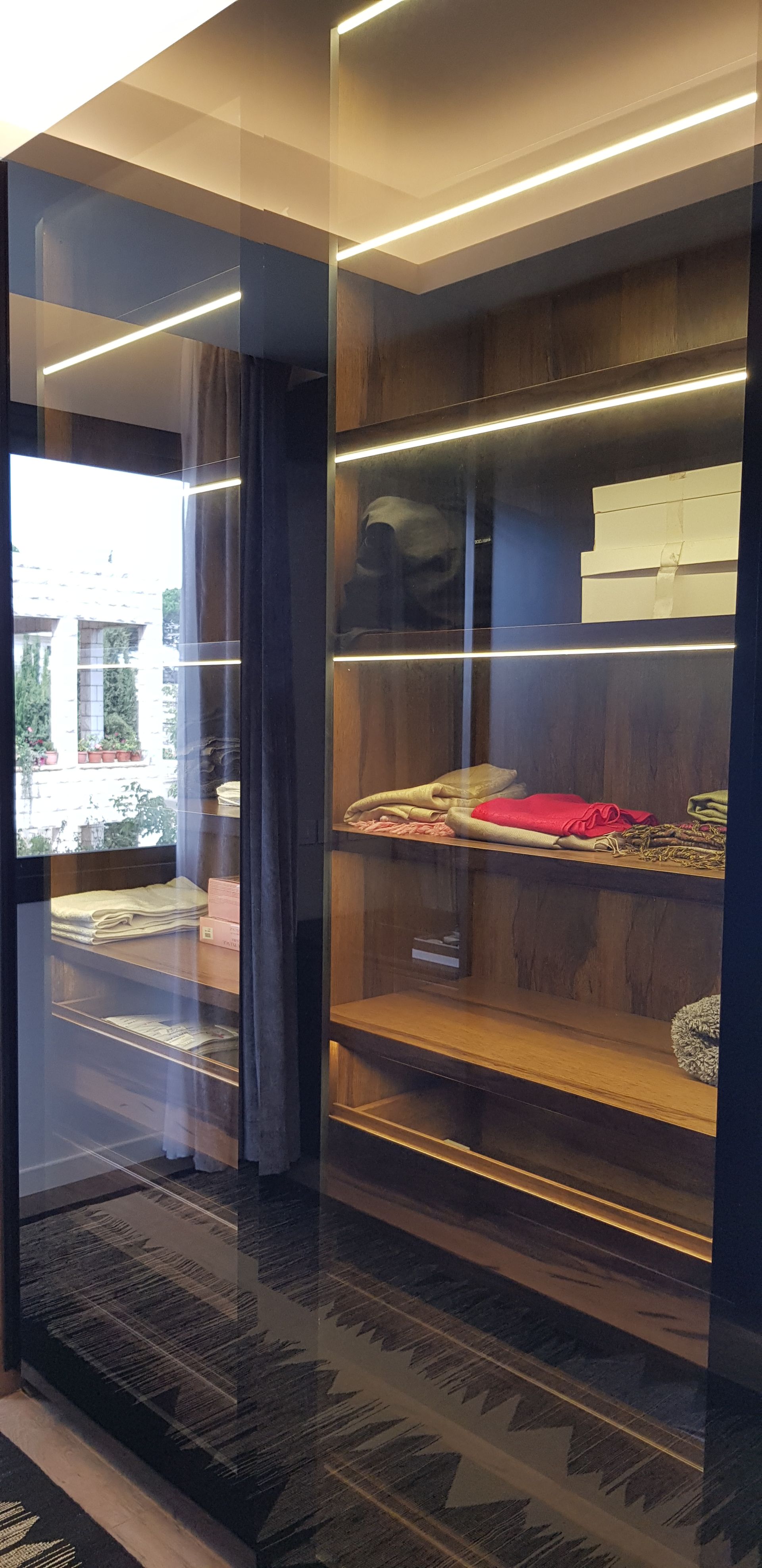 A modern walk-in closet with wooden shelves, glass doors, and strip lighting. Items such as clothing and accessories are stored inside.