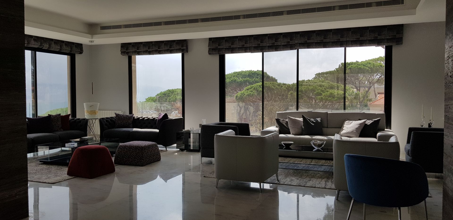 A modern living room with large windows overlooking a landscape. Furnishings include sofas and armchairs in neutral colors.