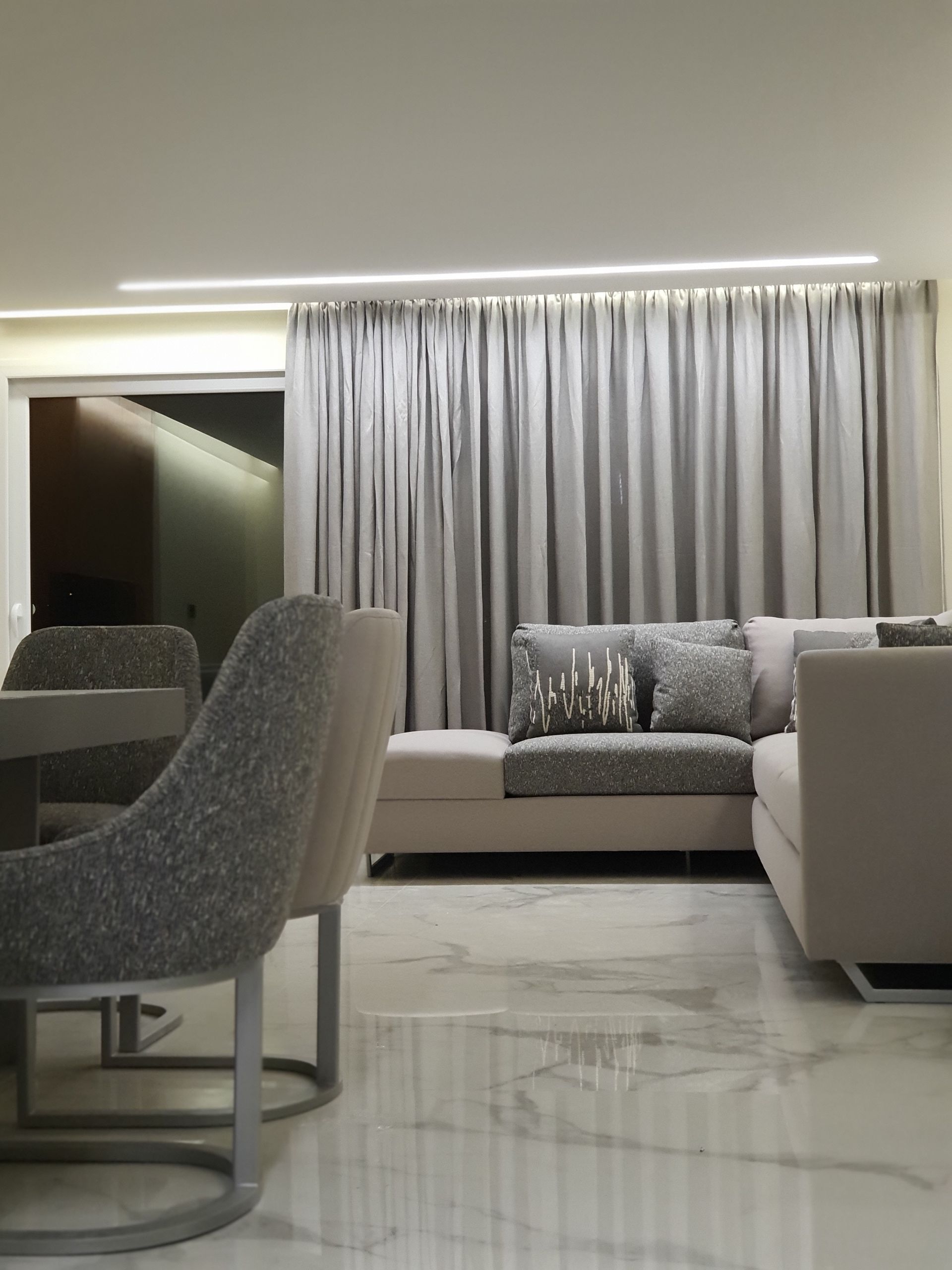 Modern living room with gray sofa, curtains, and marble floor. Dining table with chairs on the left.