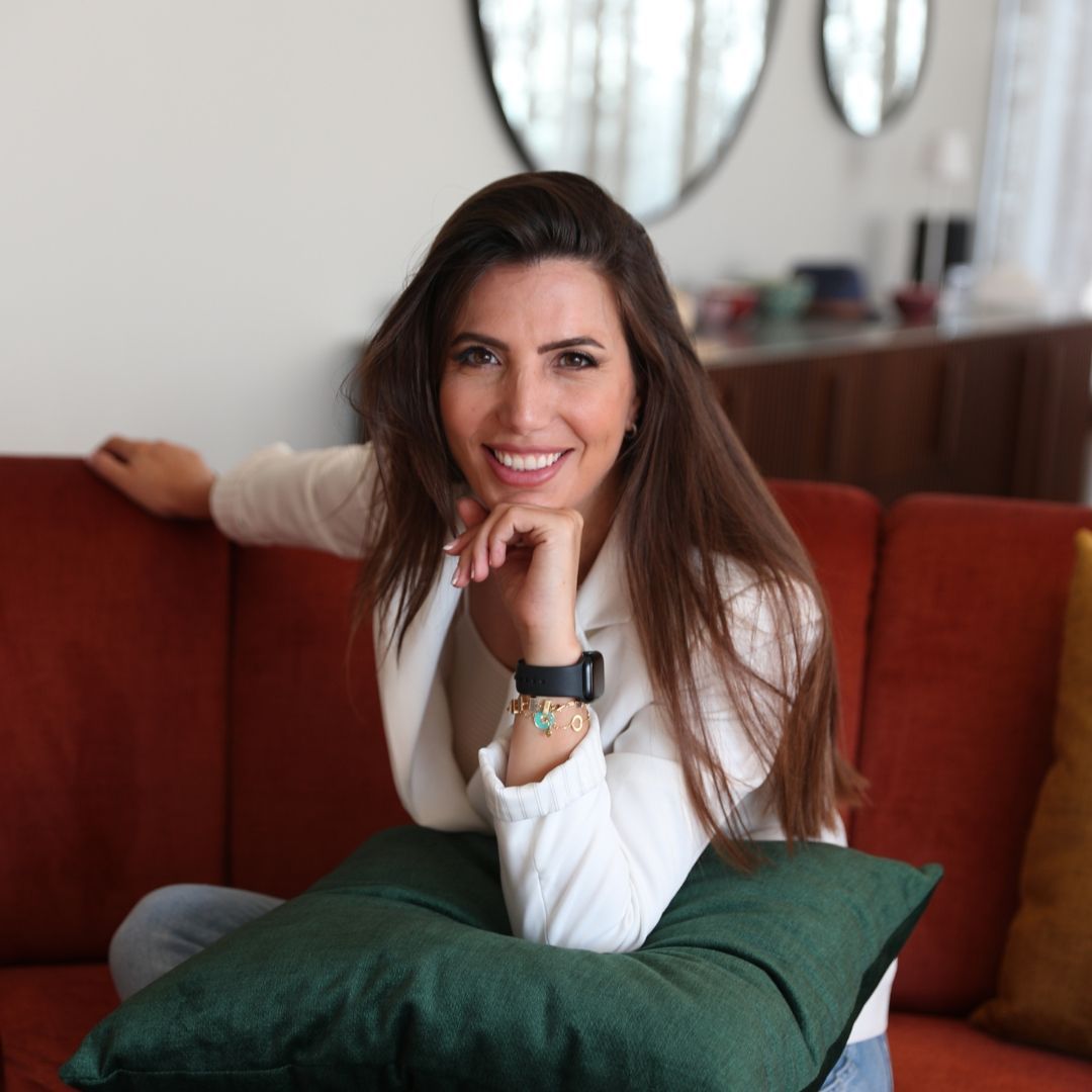 Woman with long brown hair smiles while sitting on a red couch, resting chin on hand.