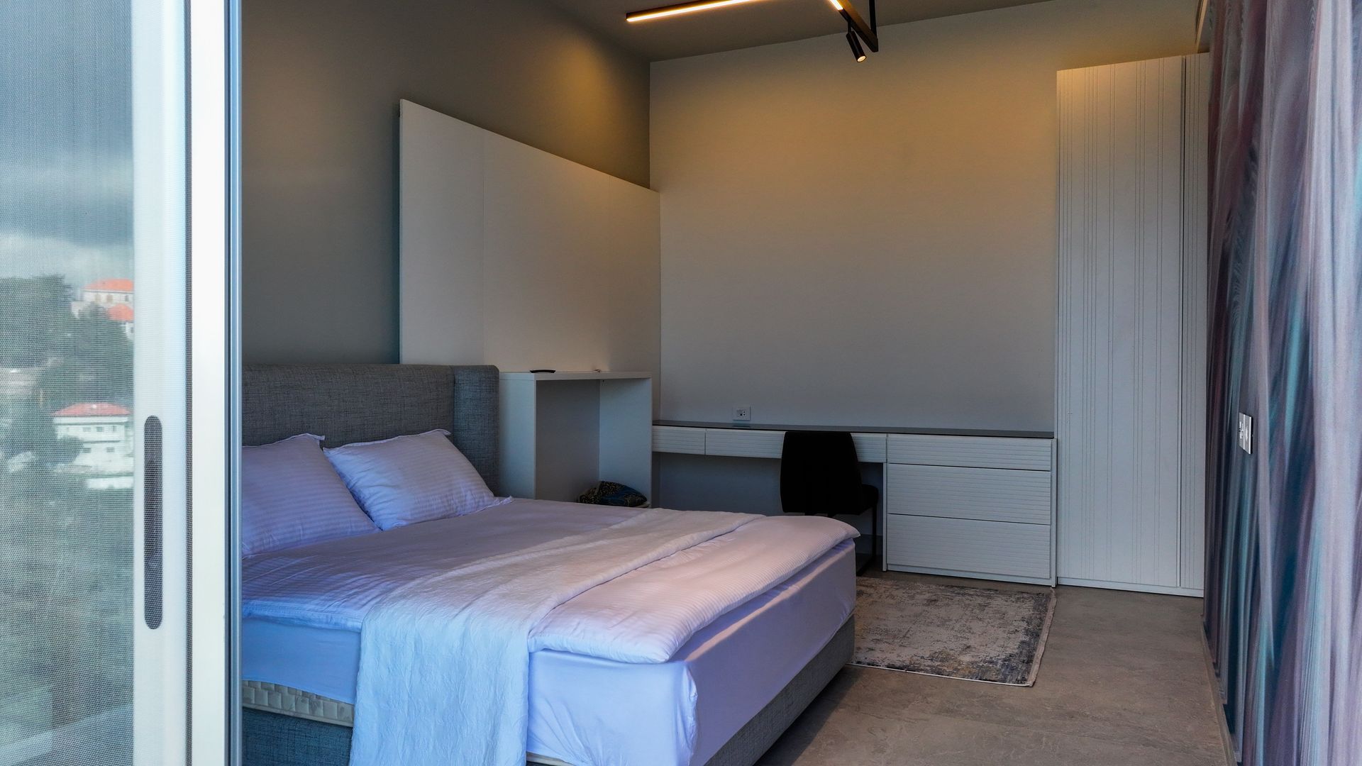 Bedroom with bed, desk, and sliding door to a balcony; mostly grey and white with light purple bedding.