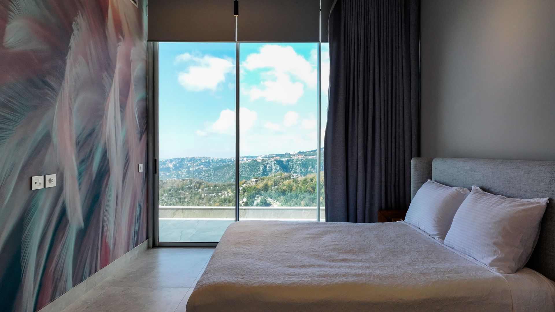Bedroom with large window overlooking a landscape; bed with white pillows, patterned closet, and grey curtains.