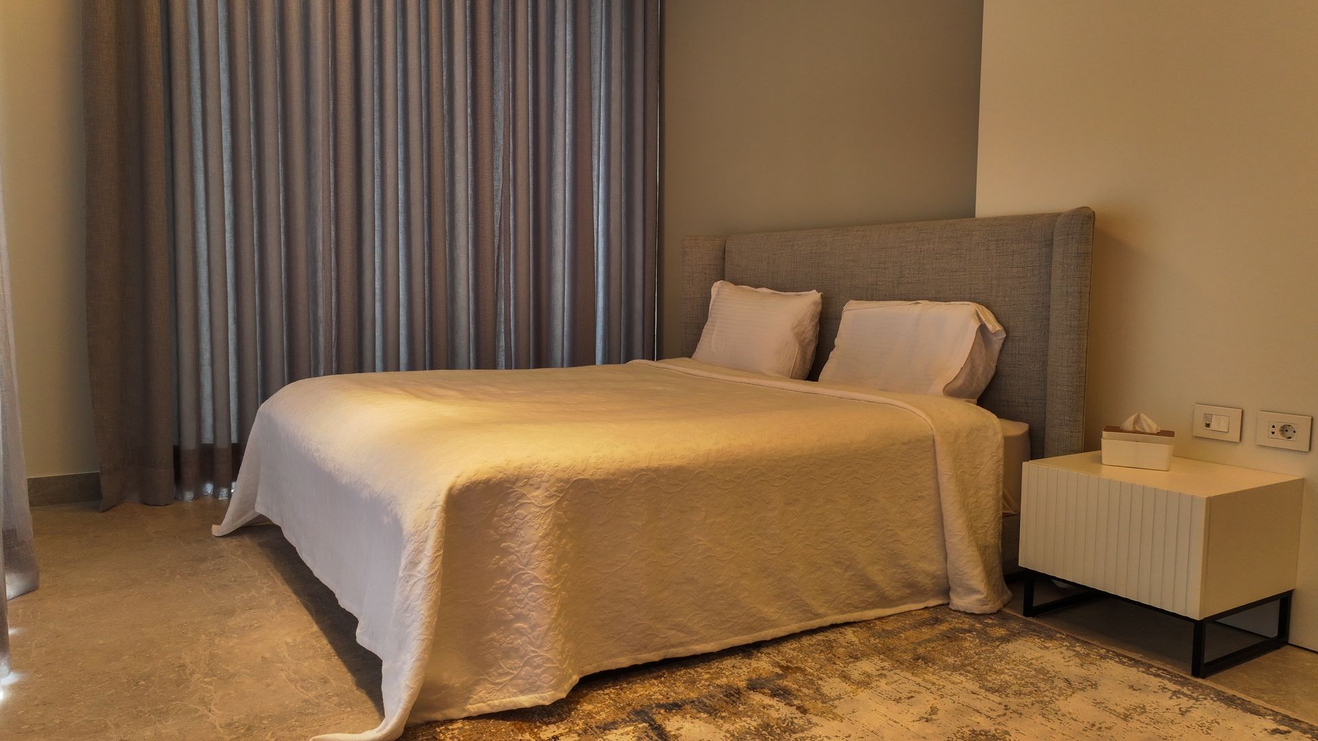 Bedroom with a bed, nightstand, curtains, and rug. The bed has white linens and a gray headboard.